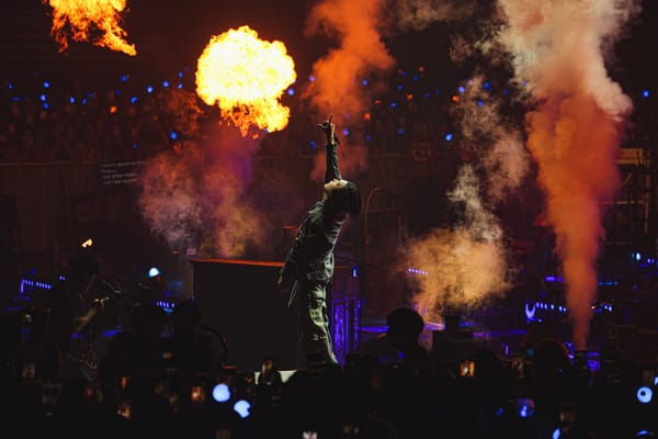 Suga on stage, raising his hand in the air as his head is thrown back, surrounded by flames and ARMY lightsticks.